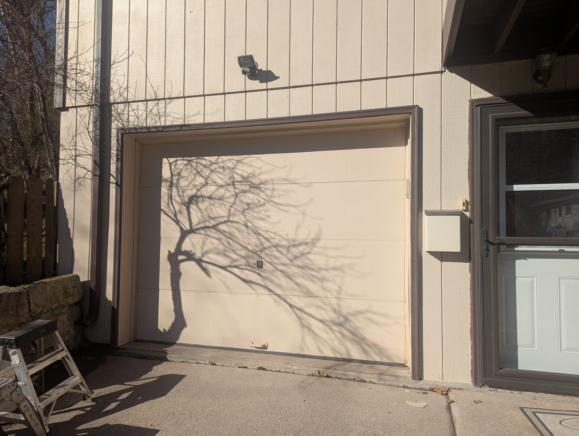 Beige garage door on a house wall with tree shadows and a small light fixture above it.