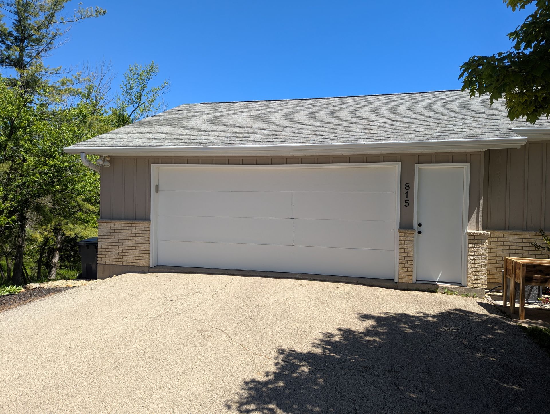 White garage door on a beige house with a gravel driveway, trees, and a bright blue sky.