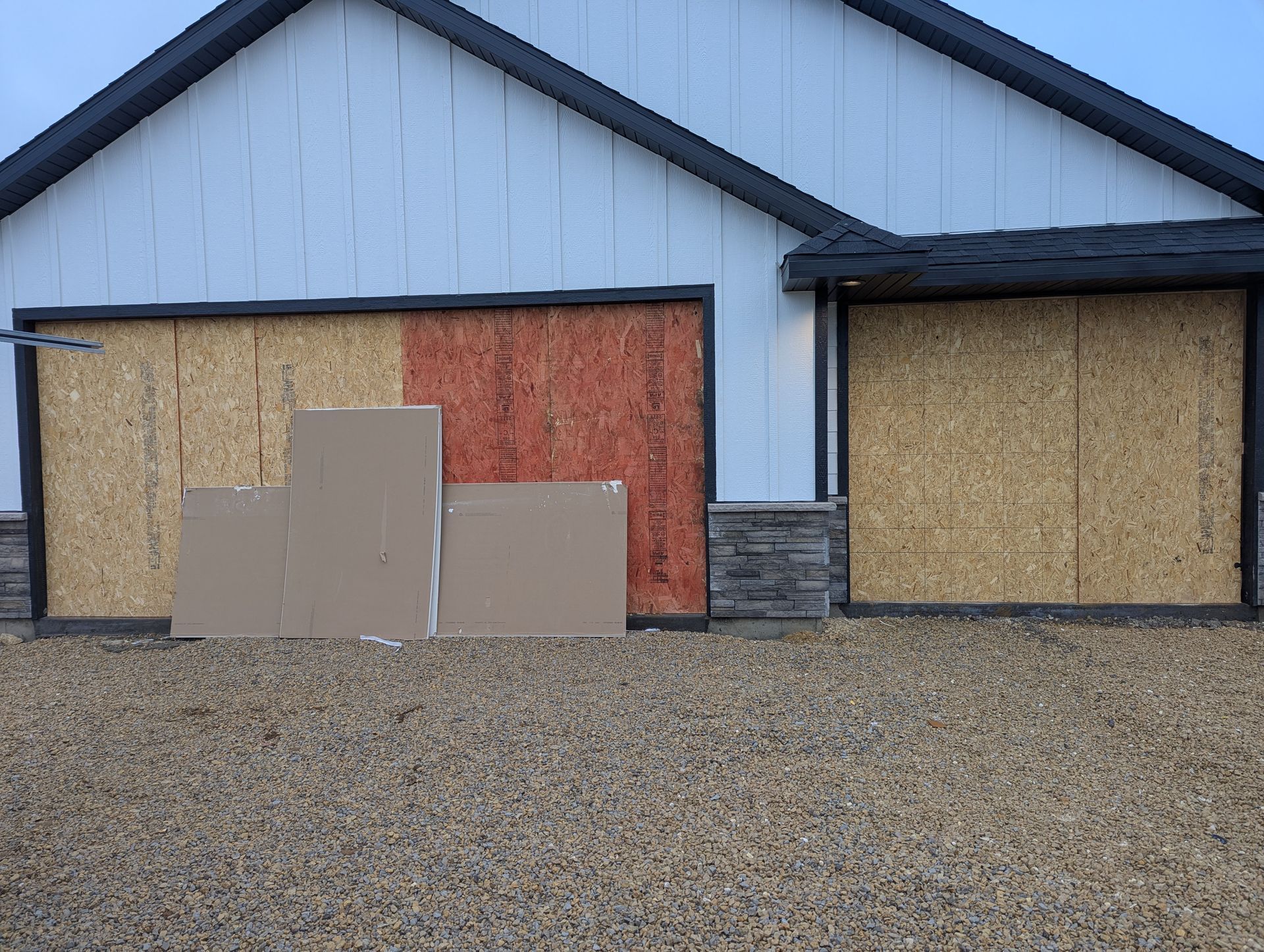 House exterior with boarded-up windows and doors, cardboard panels leaning against the front wall, gravel yard