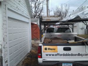 Pickup truck parked in a narrow driveway beside a garage, carrying a ladder and other tools on the back.