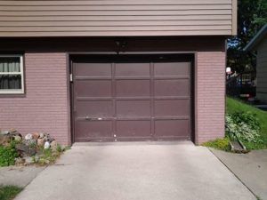 Brown garage door beneath beige siding, with driveway and small plants on both sides