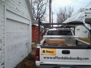 Pickup truck with ladder rack parked beside a white garage, carrying boxes and equipment.