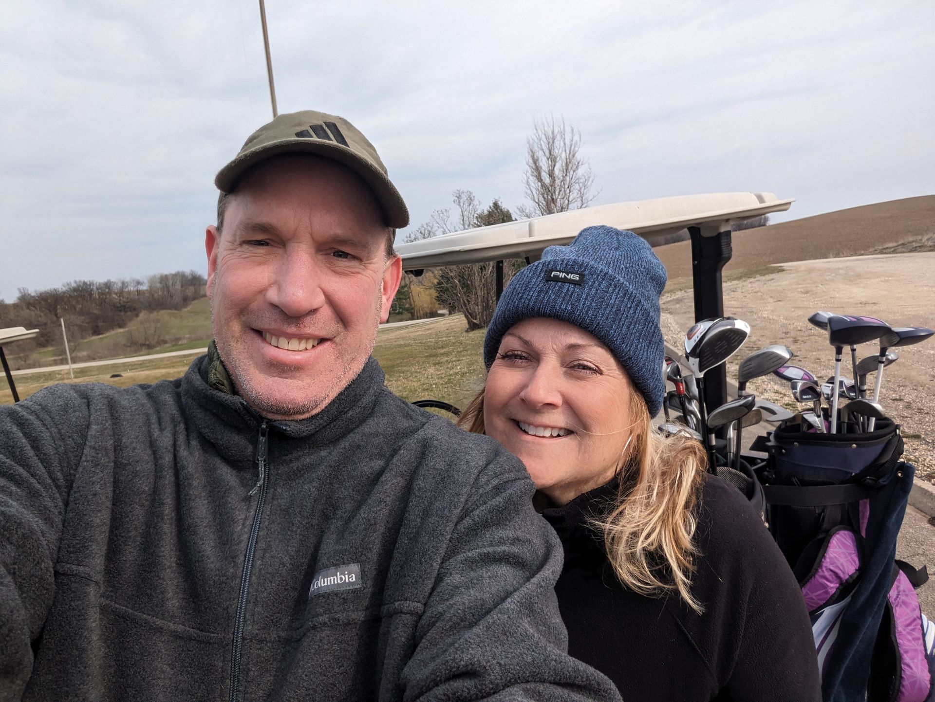 Two smiling people in outdoor winter gear pose beside golf carts on a cloudy day.