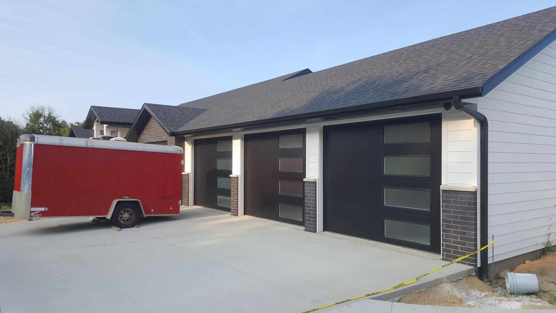 Red trailer parked beside a white brick building with dark garage doors and a driveway under construction