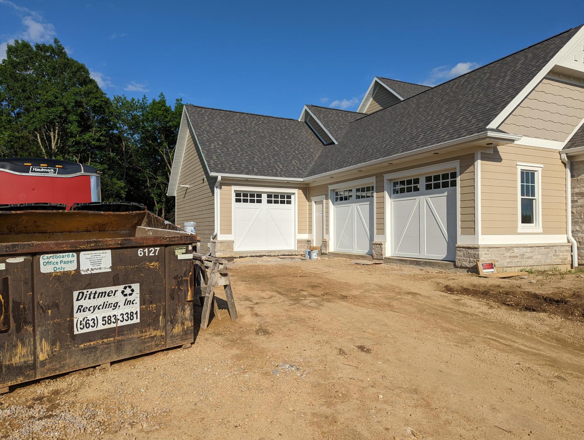 Construction dumpster beside a newly built beige house with garage doors and a dirt driveway under blue sky
