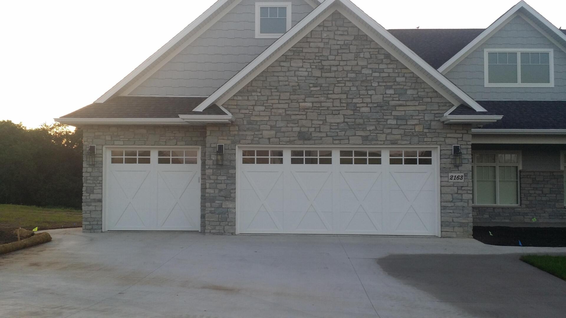 Stone house exterior with two white garage doors and a wide concrete driveway at dusk