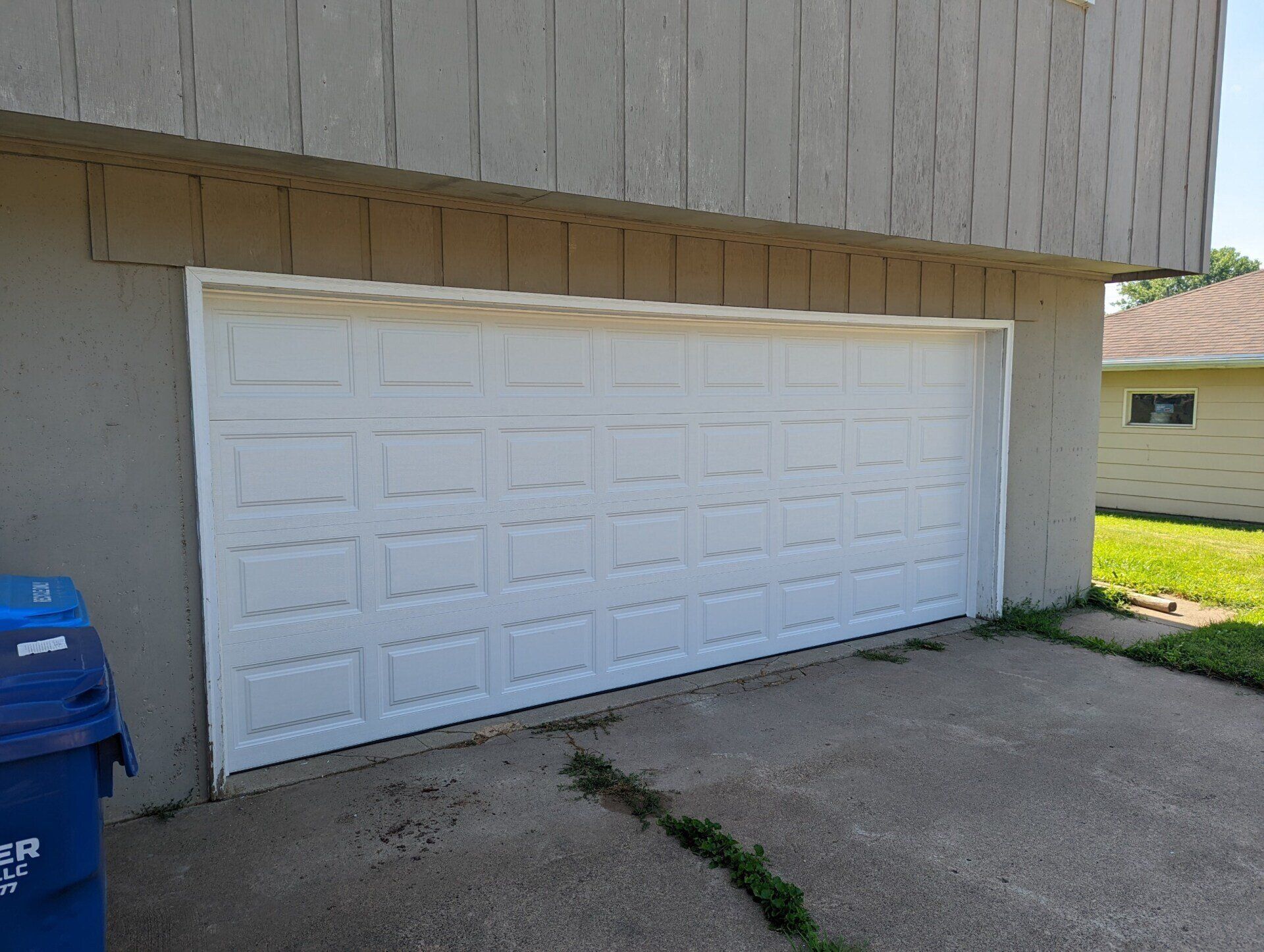 White garage door on the side of a beige building beside a driveway and blue recycling bin