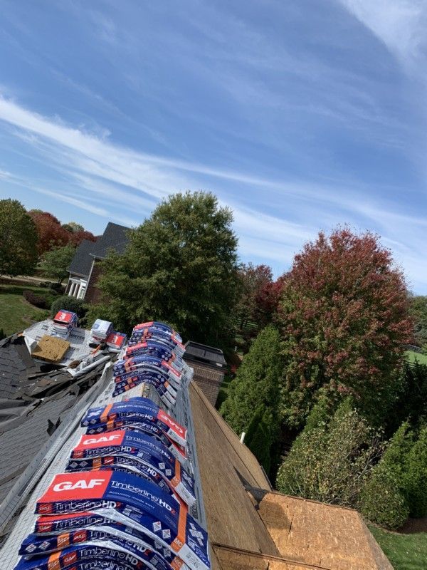 Stacked roof tiles on a house roof beside a tree-lined suburban street under a blue sky.