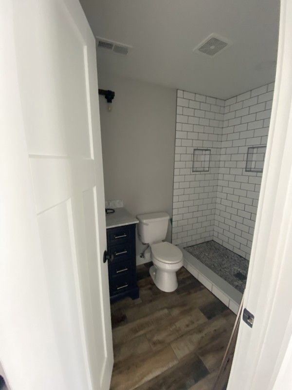 Small bathroom with white subway tile shower, dark vanity, toilet, and wood-look floor.