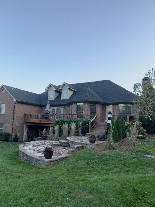 Large brick suburban house with dark roof, arched stone patio, and a grassy front lawn.