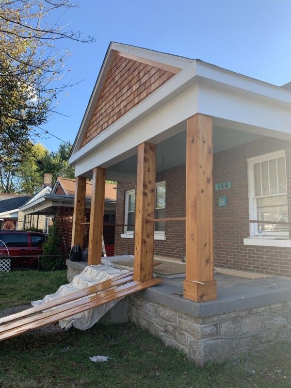 Front porch under construction with wooden posts, white trim, and stacked lumber on the steps