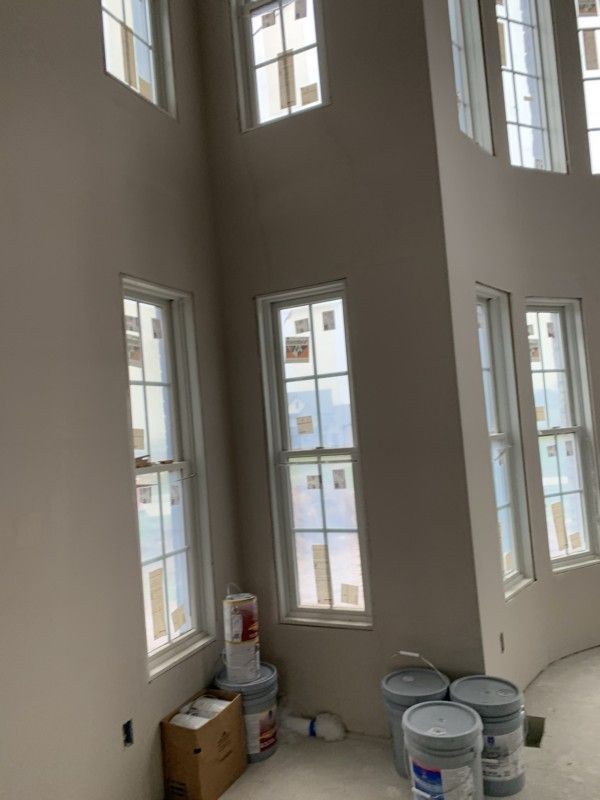 Bright, empty room with tall windows, unfinished walls, and paint buckets and boxes on the floor.