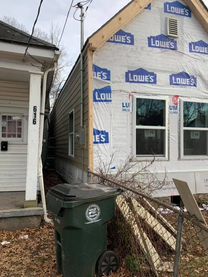 Narrow gap between houses with a green trash bin and a house wrapped in Lowe's branded siding under construction