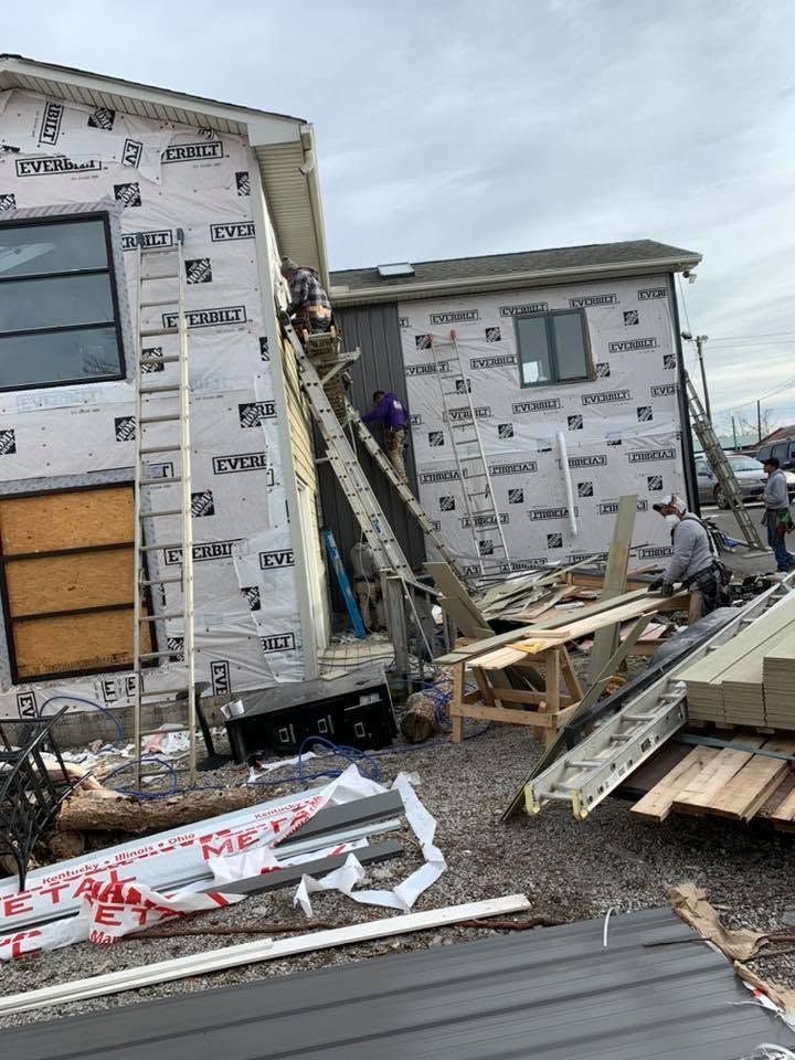 Damaged house under construction with broken siding, debris, and workers at a collapsed stairway.