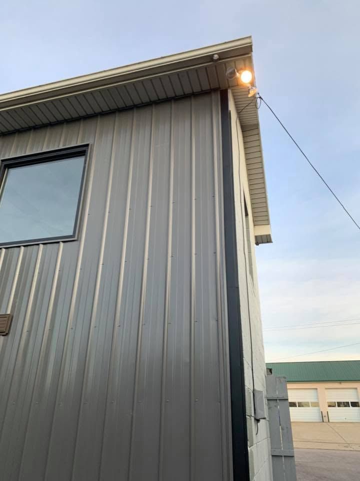Gray metal-sided building corner with a lit eave light and a window, seen against the evening sky