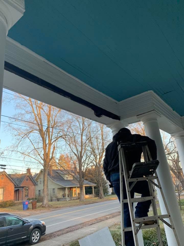 Person on a ladder painting a porch ceiling blue and white, with a street and houses in the background