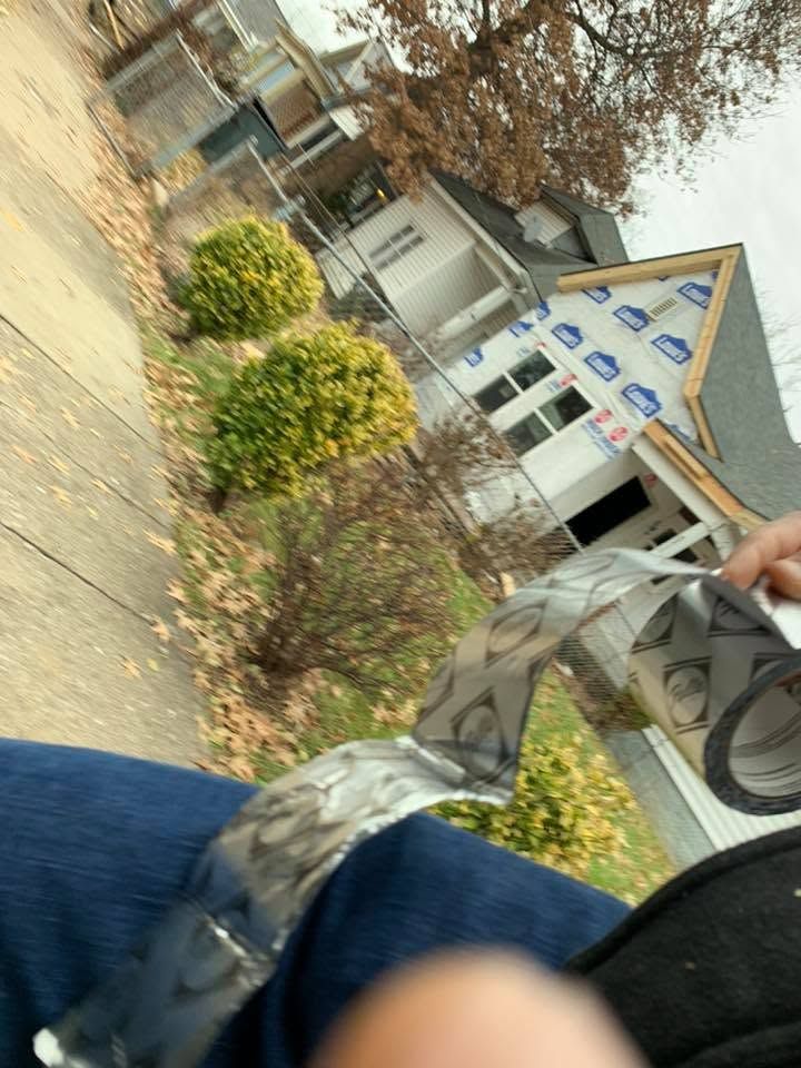 Person walking past a building with bushes and trees on a sidewalk, carrying a bag