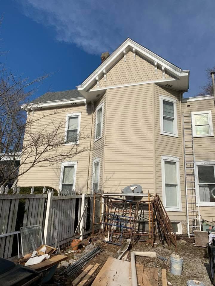 Damaged beige house exterior with broken porch, debris, and a wooden fence under a blue sky