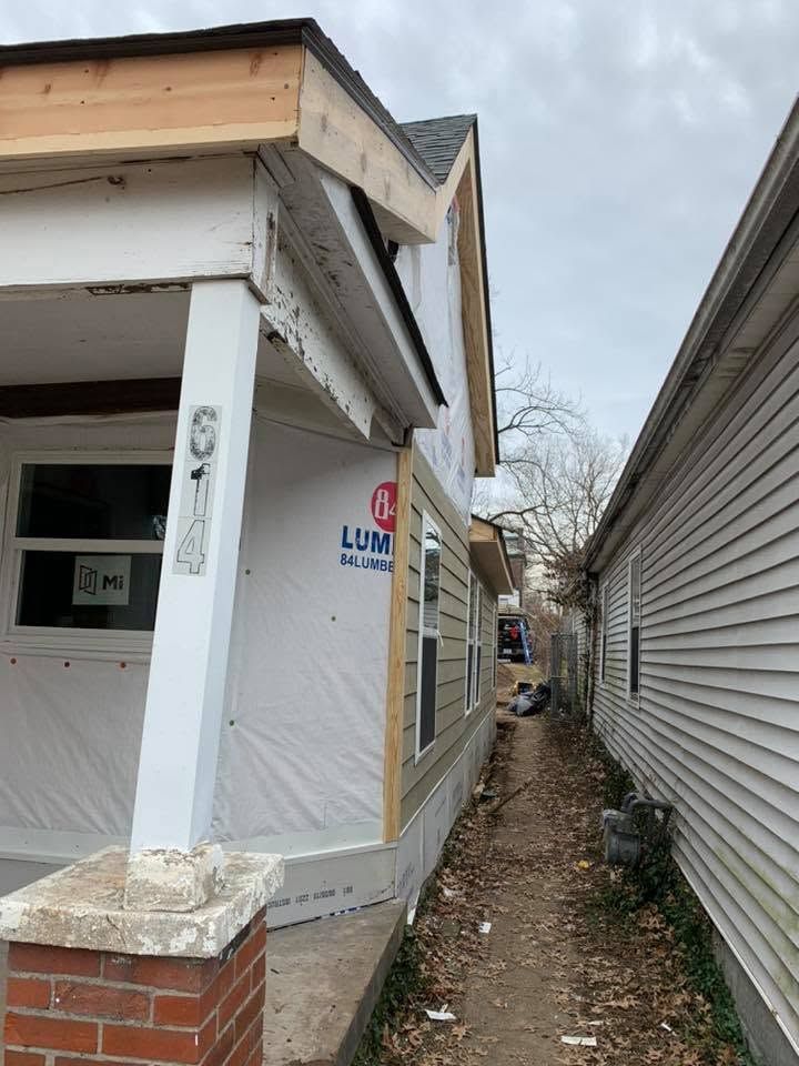 Damaged porch beside a narrow alley between houses, with exposed framing and construction wrap
