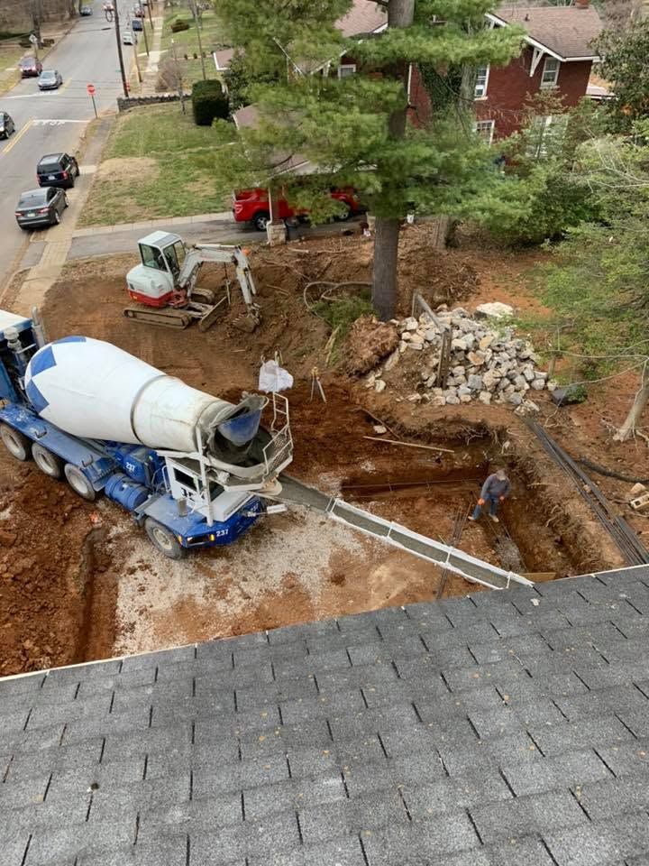 Concrete truck pumping into a muddy construction trench beside a house and large tree