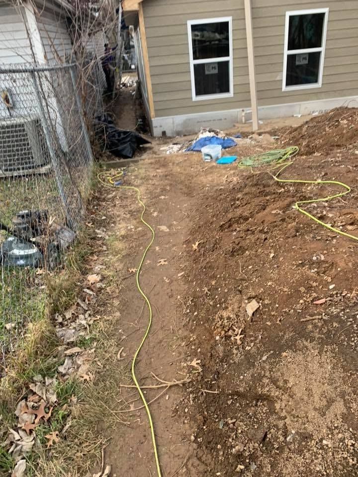 Narrow dirt trench beside a house, with a hose or cable running along the ground near the foundation.
