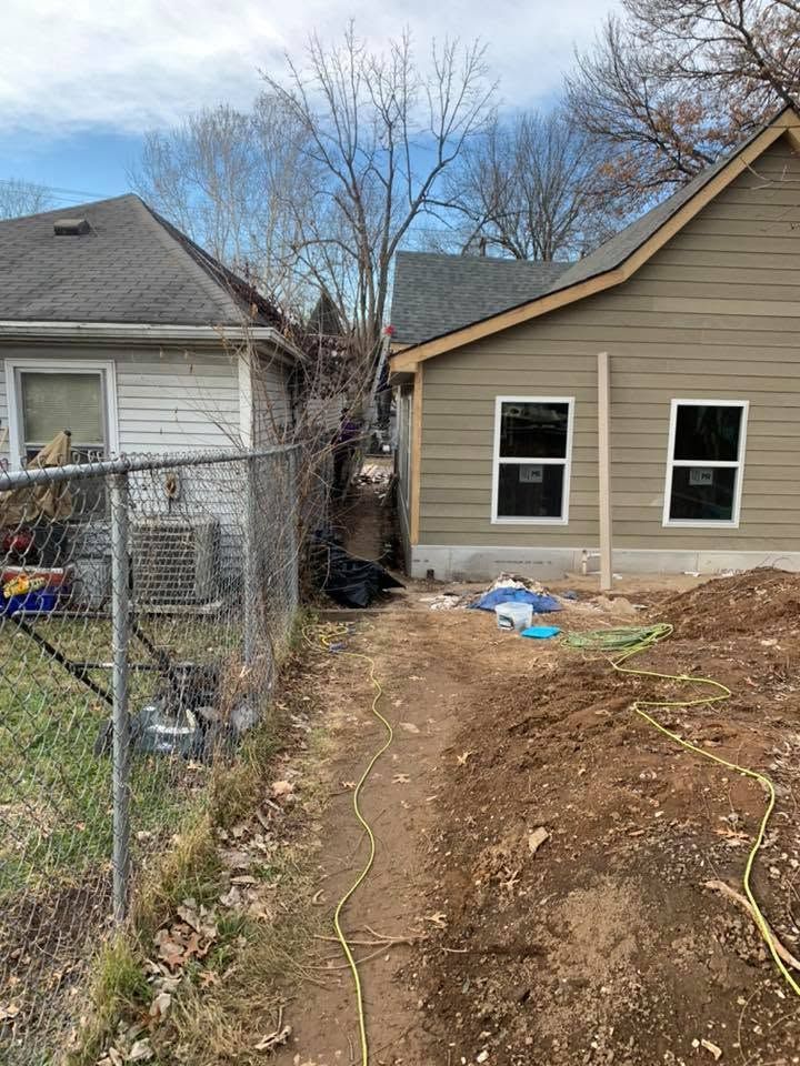 Narrow dirt path between a chain-link fence and tan house, with yard debris and a hose along the ground.