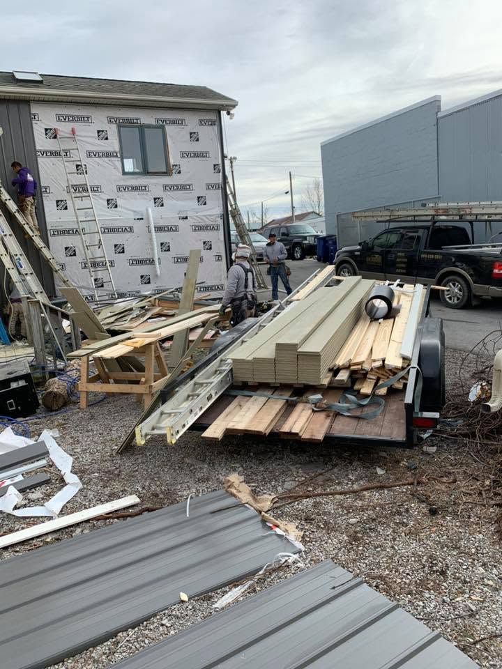 Construction site with house siding, lumber, and debris piled beside a building and parked pickup truck.