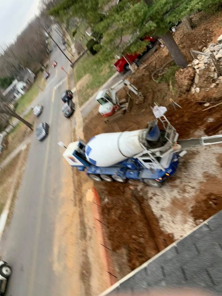 Overturned cement mixer truck on a muddy roadside beside a paved street, with cars passing nearby.
