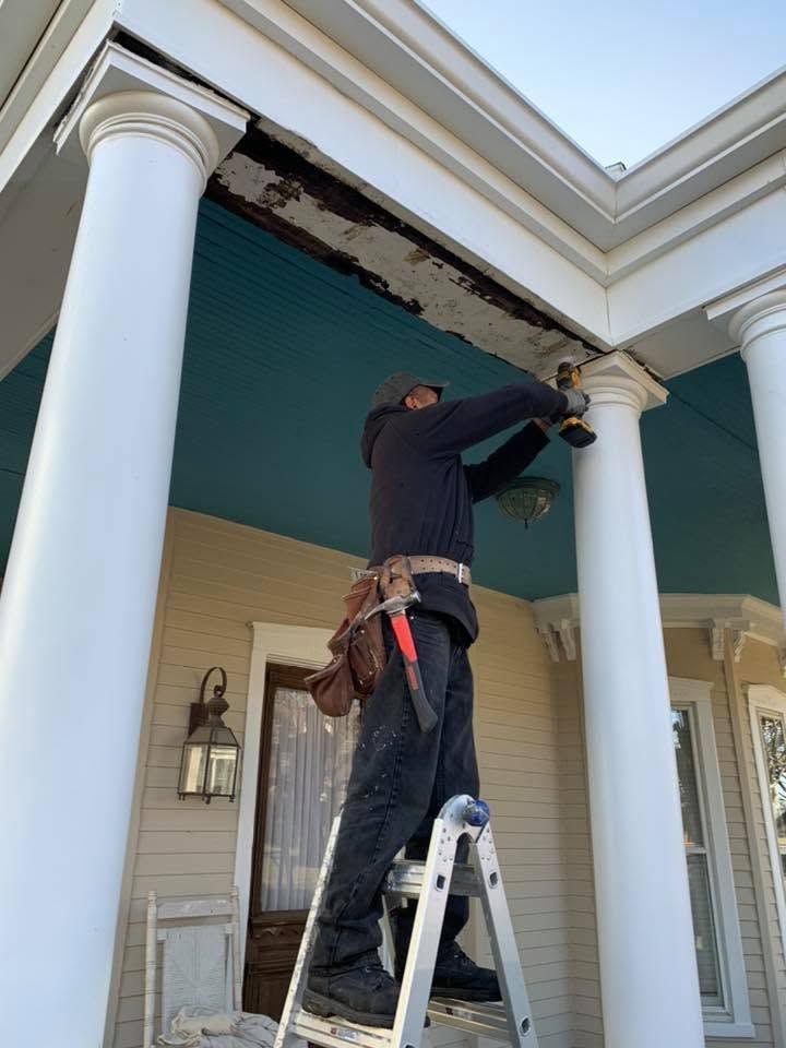 Worker on a ladder repairing peeling paint on a porch ceiling between white columns.