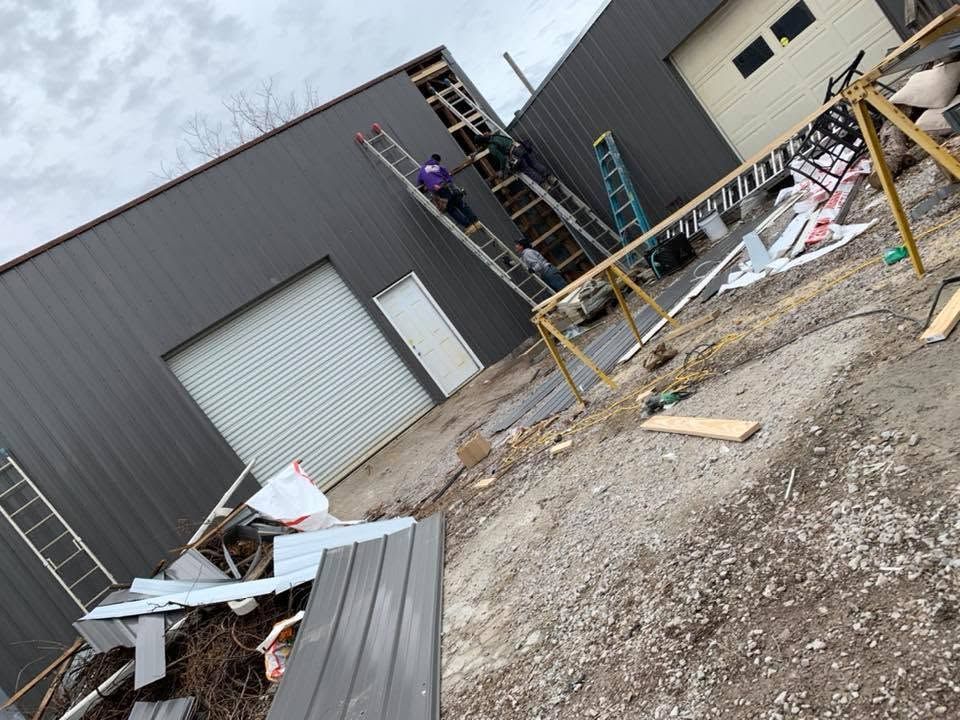 Construction site outside warehouse with debris, ladders, and two workers on scaffolding.