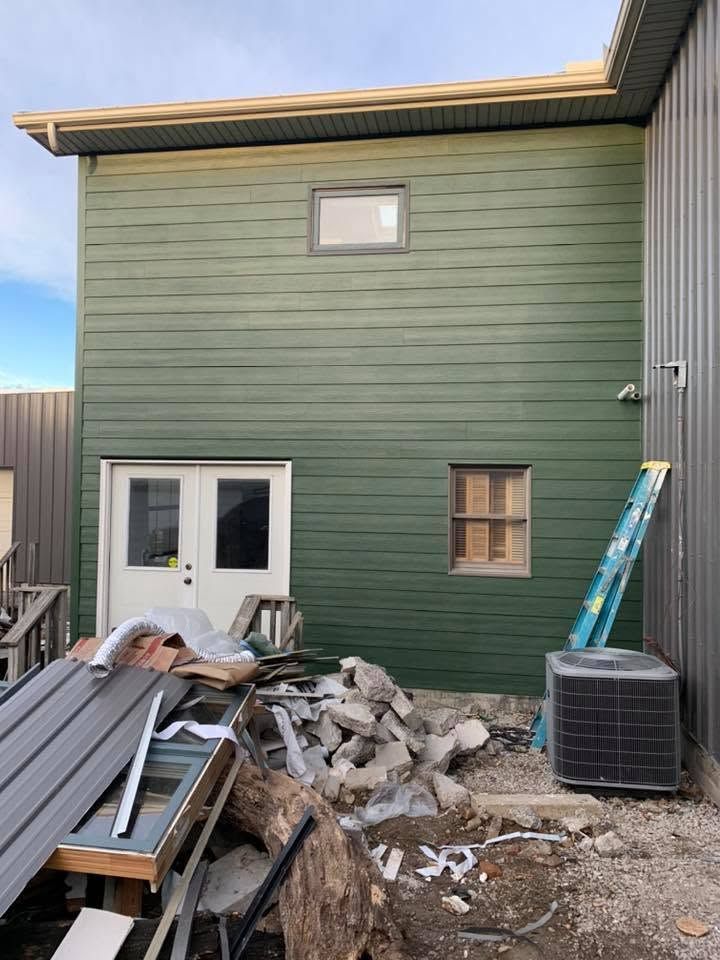 Green house exterior with white doors and window, piles of construction debris in the yard, and an AC unit on the right