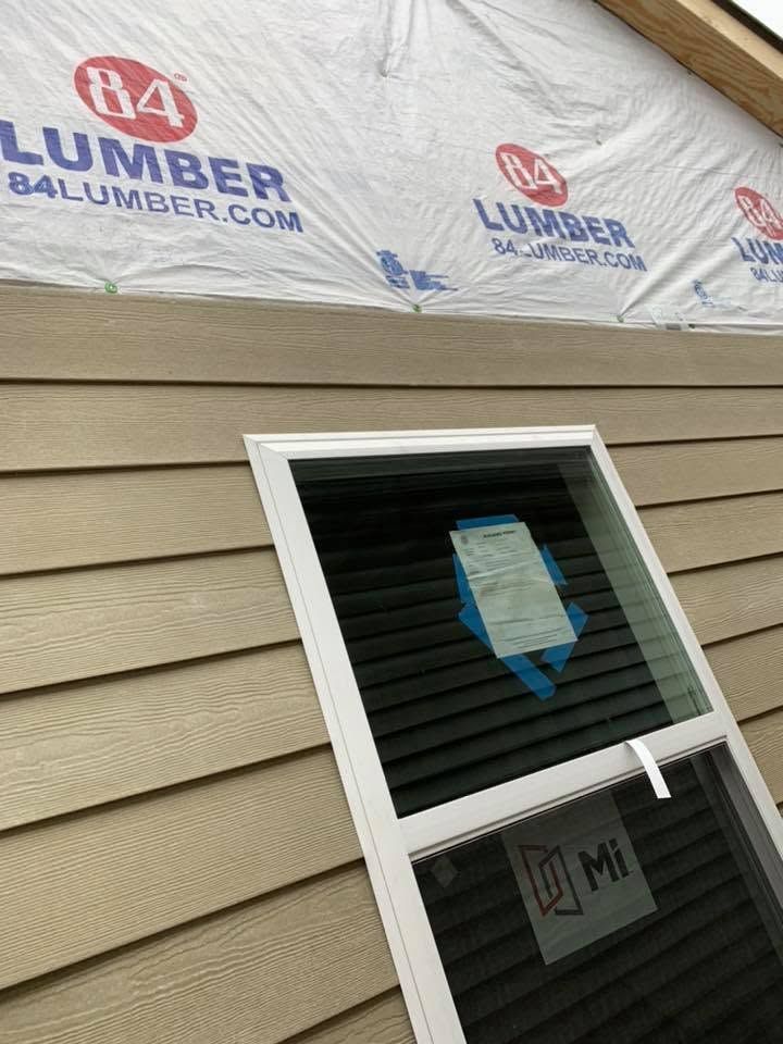 Window in beige siding with blue sticker, under roof wrap marked “84 Lumber.”