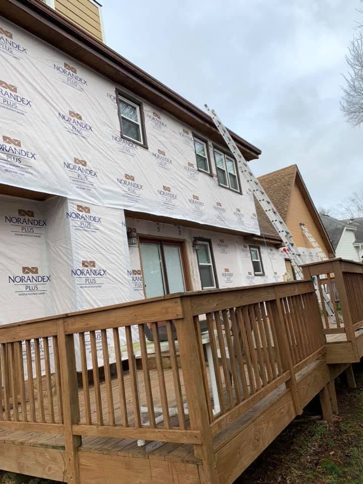 Partially renovated house wrapped in white siding paper beside a wooden deck and fence