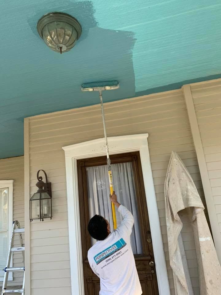 Person painting a porch ceiling blue while standing on a ladder near a door