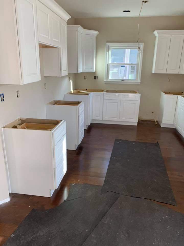 Unfinished kitchen with white cabinets, brown floor, and dark floor mats in front of the sink and cabinets