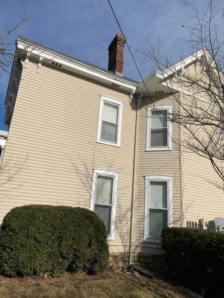 Beige two-story house with white trim, brick chimney, and trimmed bushes under a blue sky.