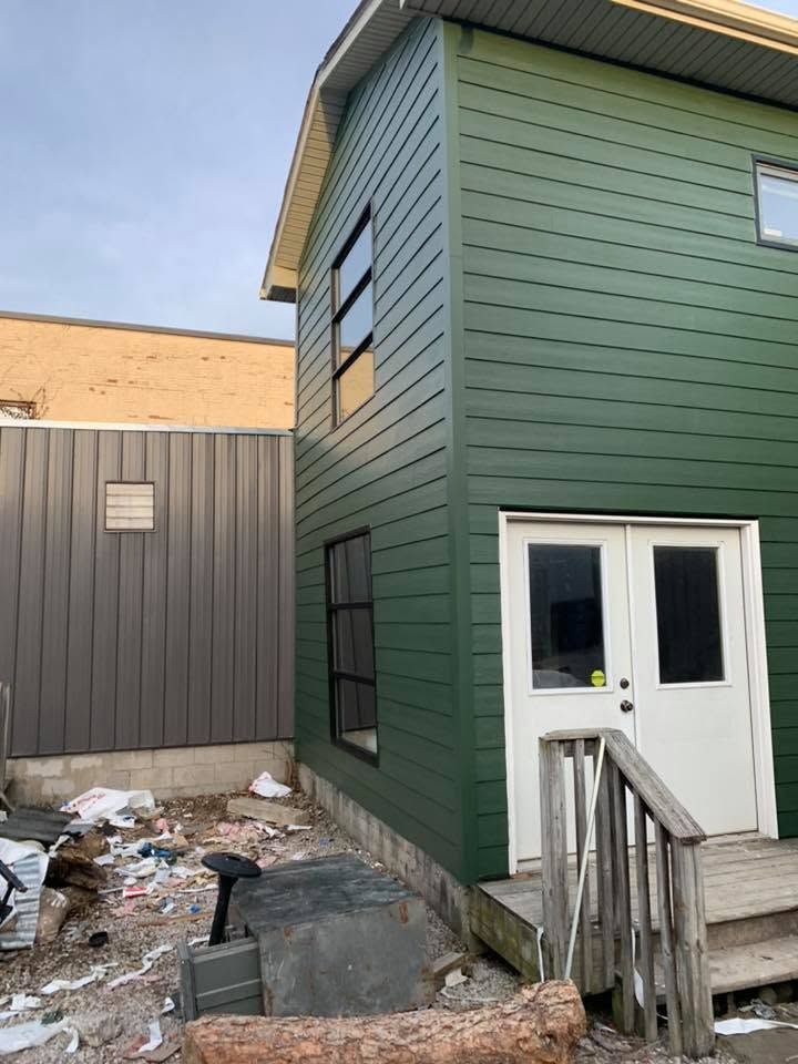 Green-sided house with white doors beside a narrow snowy alley and unfinished ground work