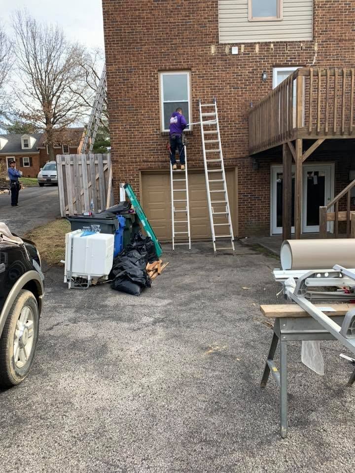 Person standing on a ladder beside a brick house, with a second ladder and cleanup materials in the driveway.