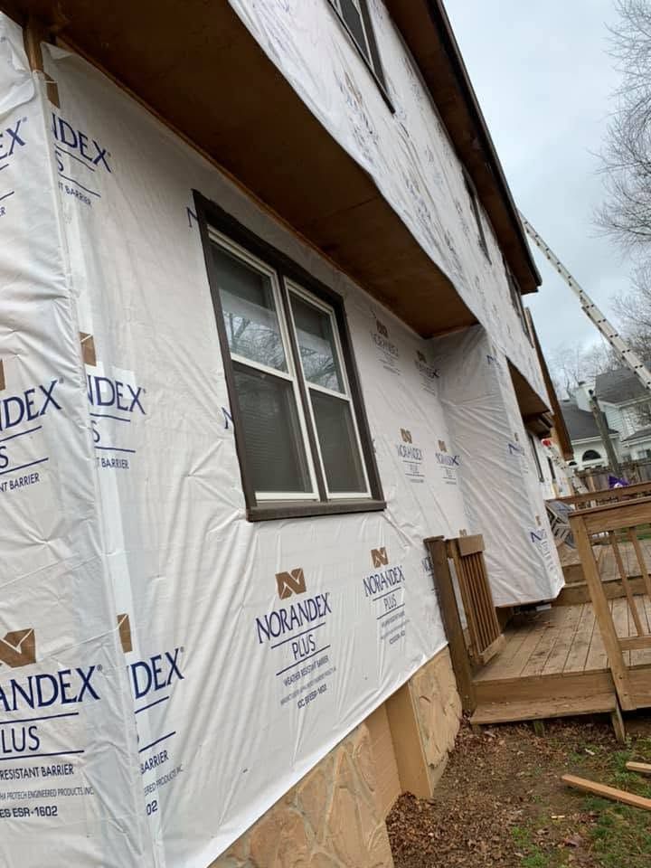 House exterior wrapped in white Tyvek housewrap during renovation, with window and wooden porch visible