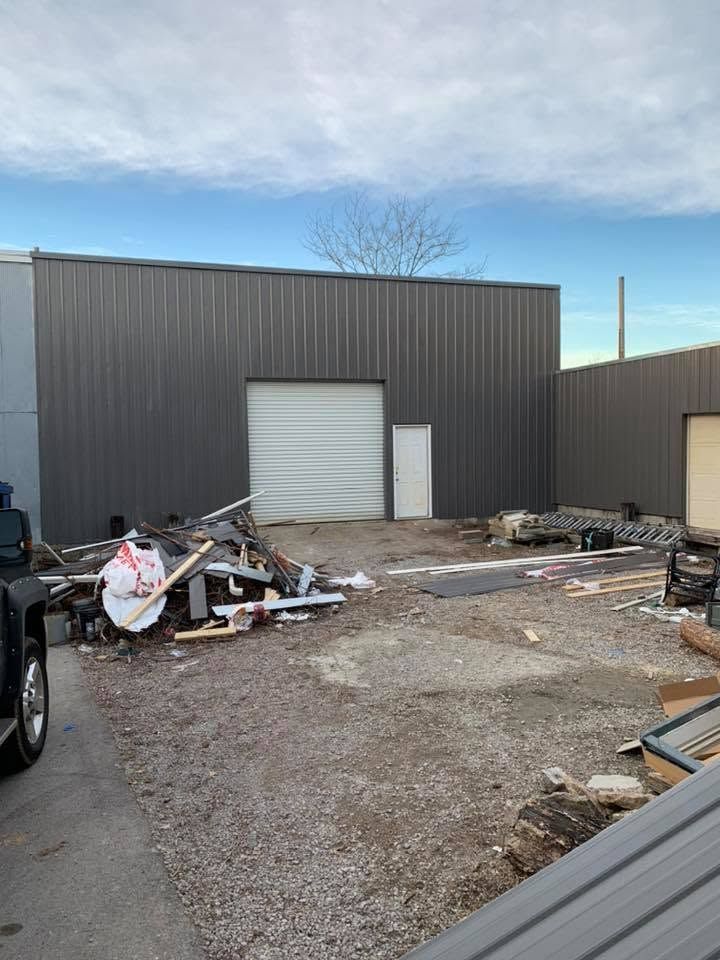 Industrial yard with a gray metal building, garage door, and piles of scrap metal and debris.