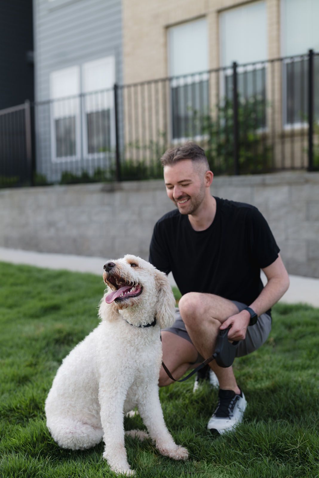 Man kneeling with a white dog on a grassy area, smiling. Buildings and a fence in the background.