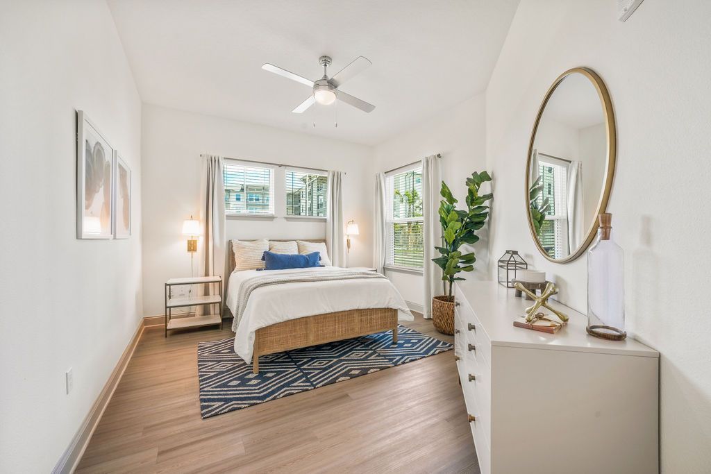 Bedroom with bed, dresser, mirror, and windows; white walls, wood floors, neutral tones.