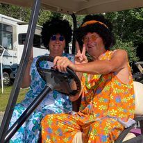 Two people in bright orange outfits and wigs sitting in a golf cart, smiling and posing with peace signs