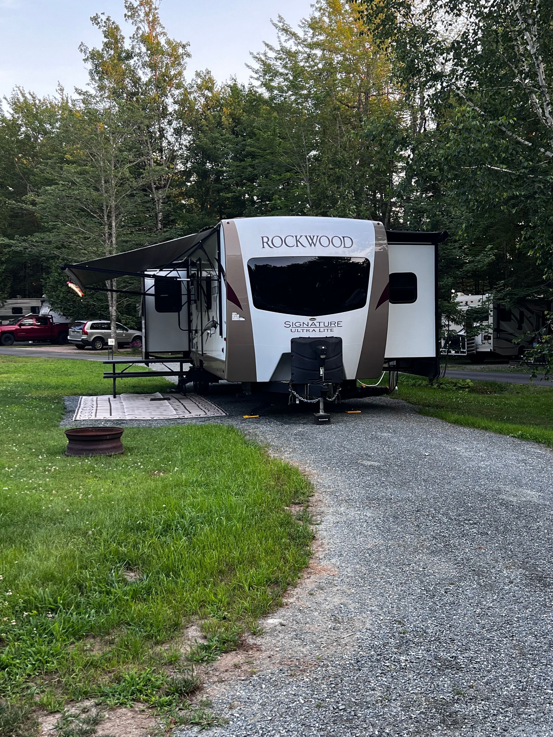White RV parked on a gravel campsite among trees, with an awning and picnic area.