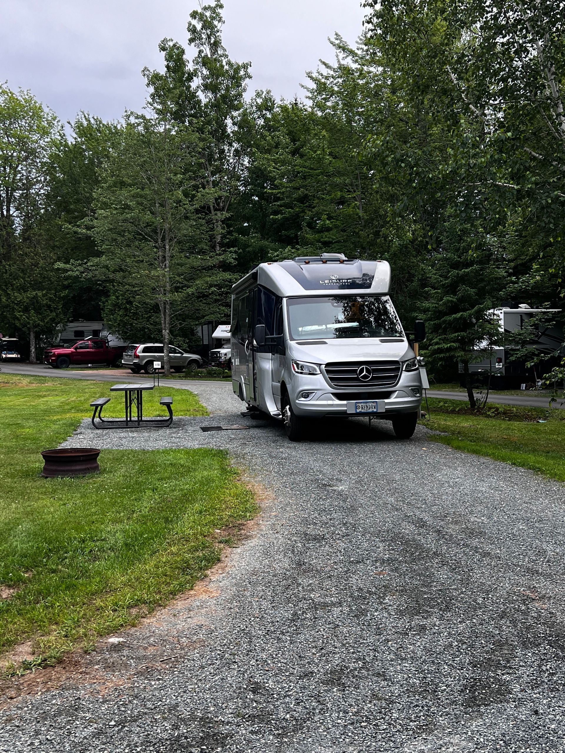 Silver RV parked on a gravel campsite surrounded by trees and grass