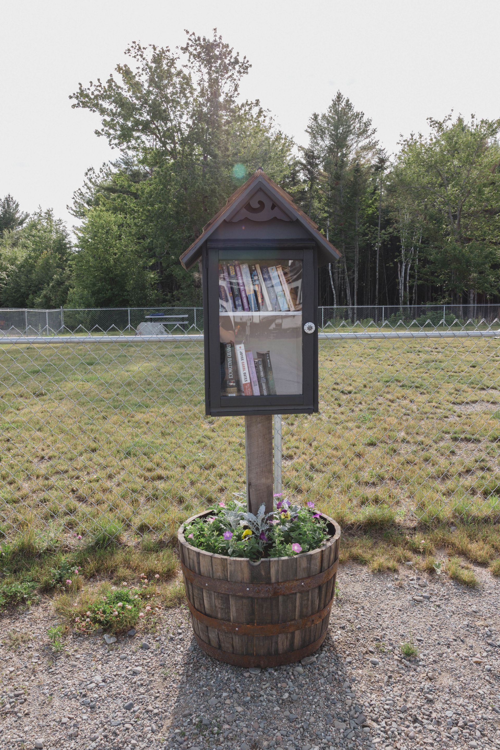 Small memorial display in a wooden planter barrel on a gravel path, with trees in the background.