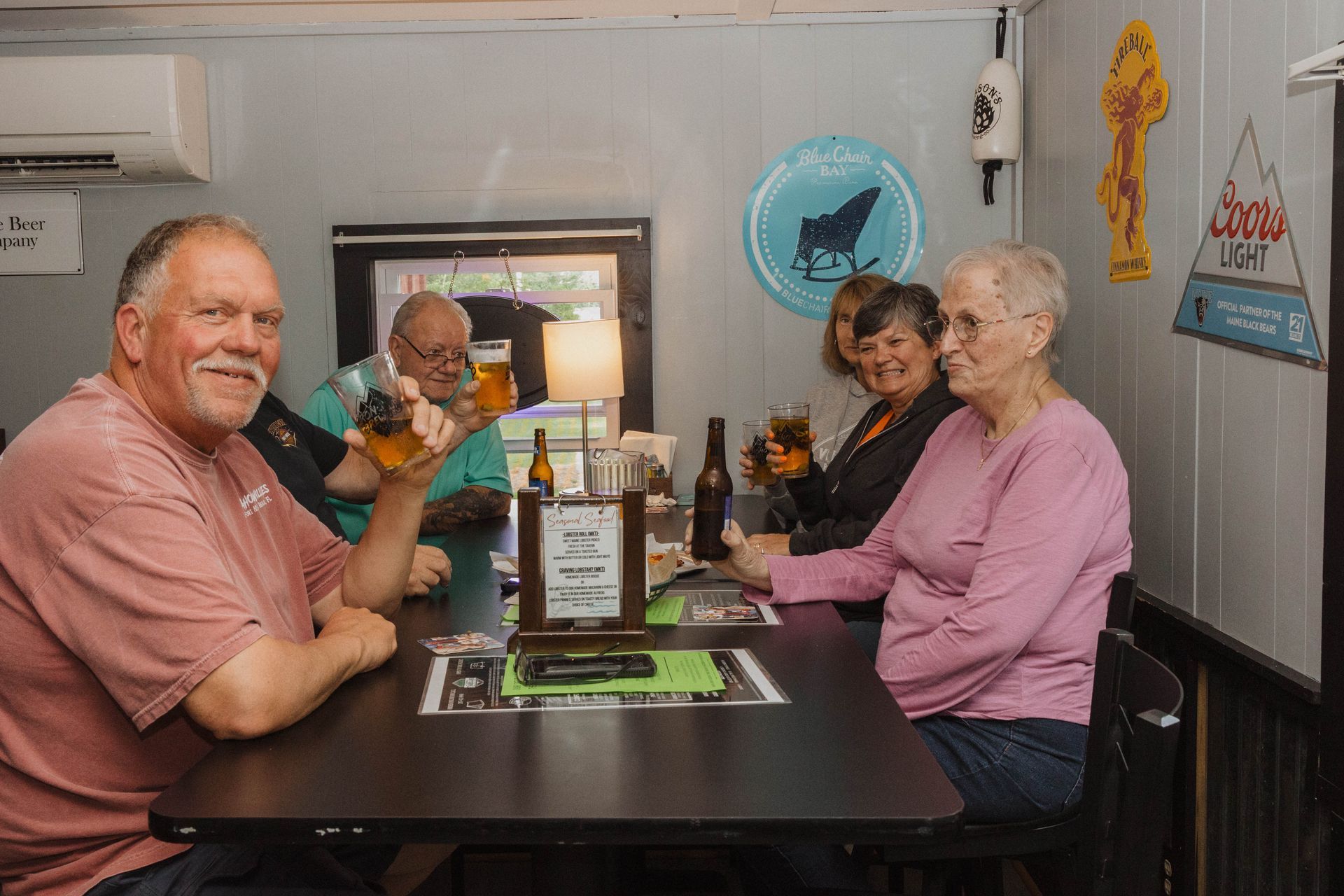 Three people seated at a diner table, smiling indoors near a window and wall decor.
