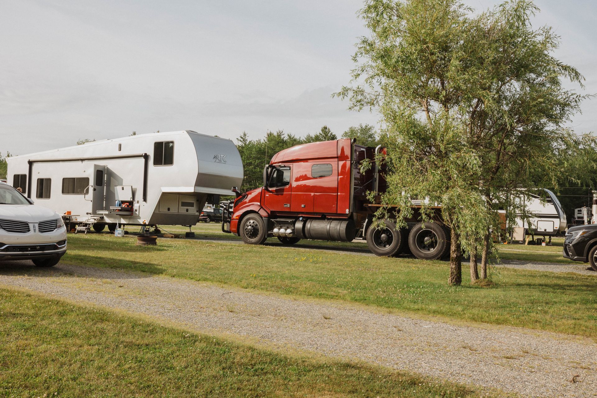 Red semi-truck and white RV parked on grass at a campground with trees and a silver car nearby