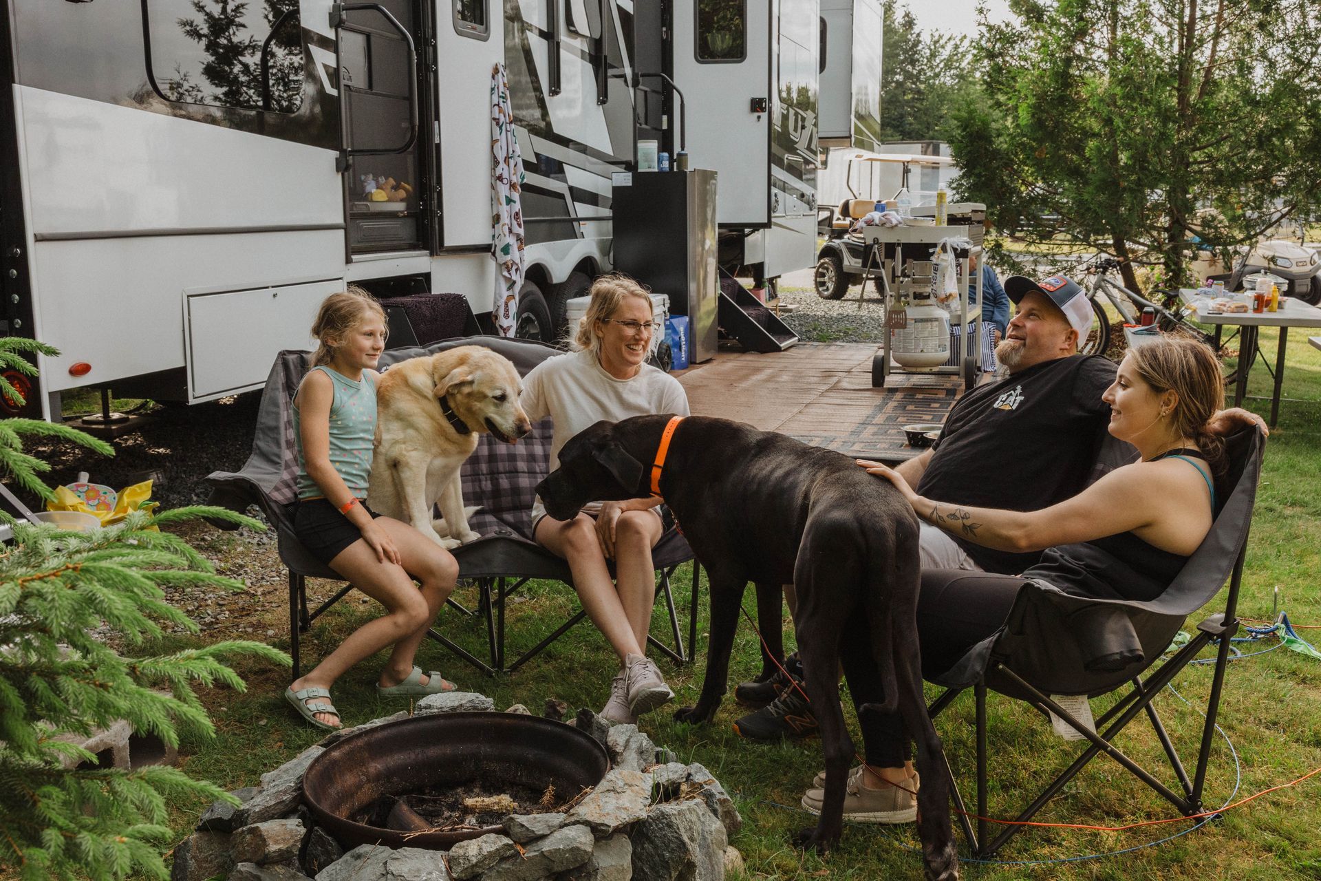 Group chatting around a campfire beside RVs, with chairs, trees, and a black dog in the foreground