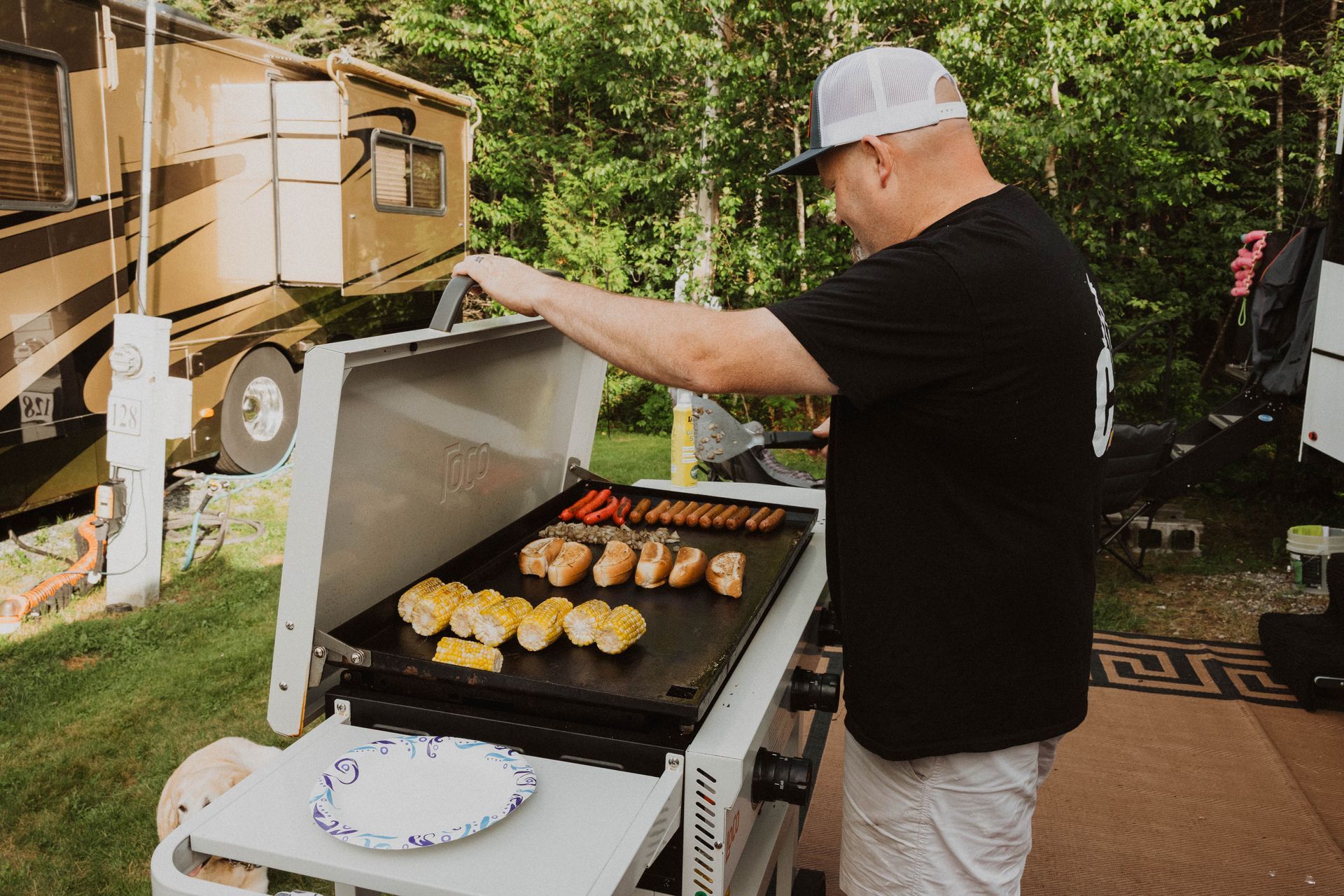 Person grilling food on an outdoor barbecue beside a tan RV in a wooded campsite.